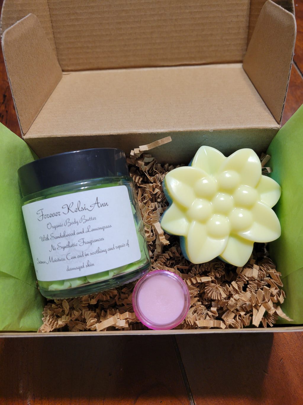 An open gift box containing a jar of Forever Kelsi Ann body butter, a bar of handcrafted soap, and a container of lip balm.