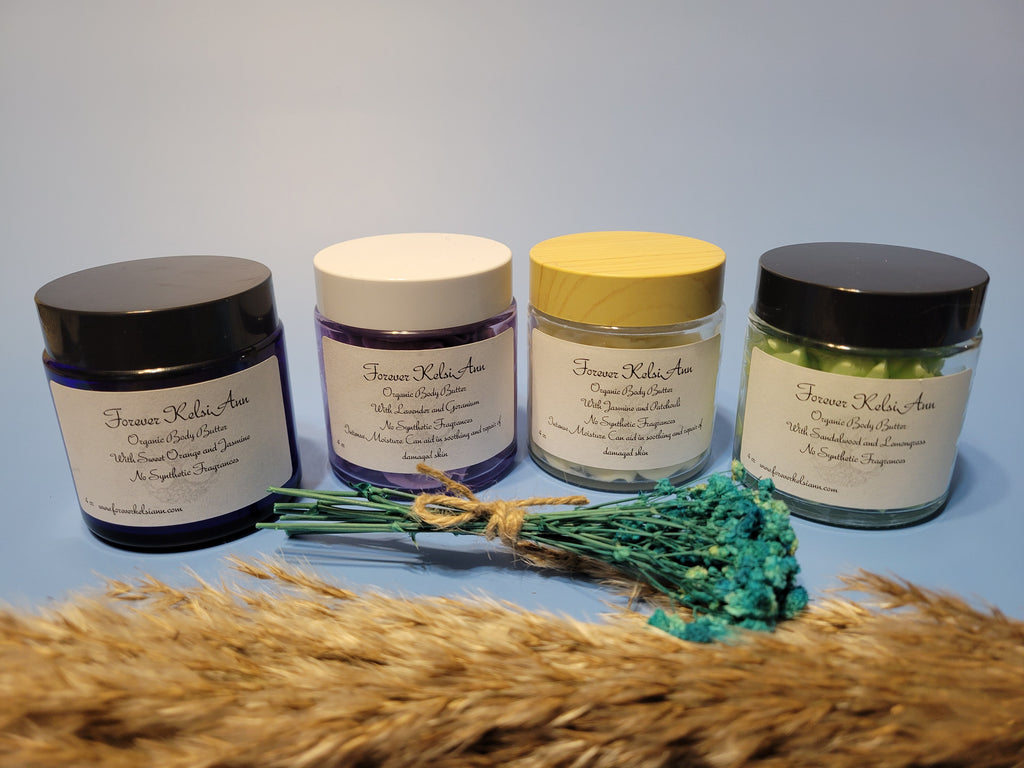 Four glass jars of assorted Forever Kelsi Ann organic body butter on a light blue surface with decorative elements.