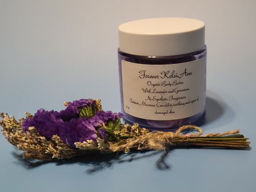 Jar of Forever Kelsi Ann organic body butter with lavender and geranium, surrounded by purple flowers on a light blue background.