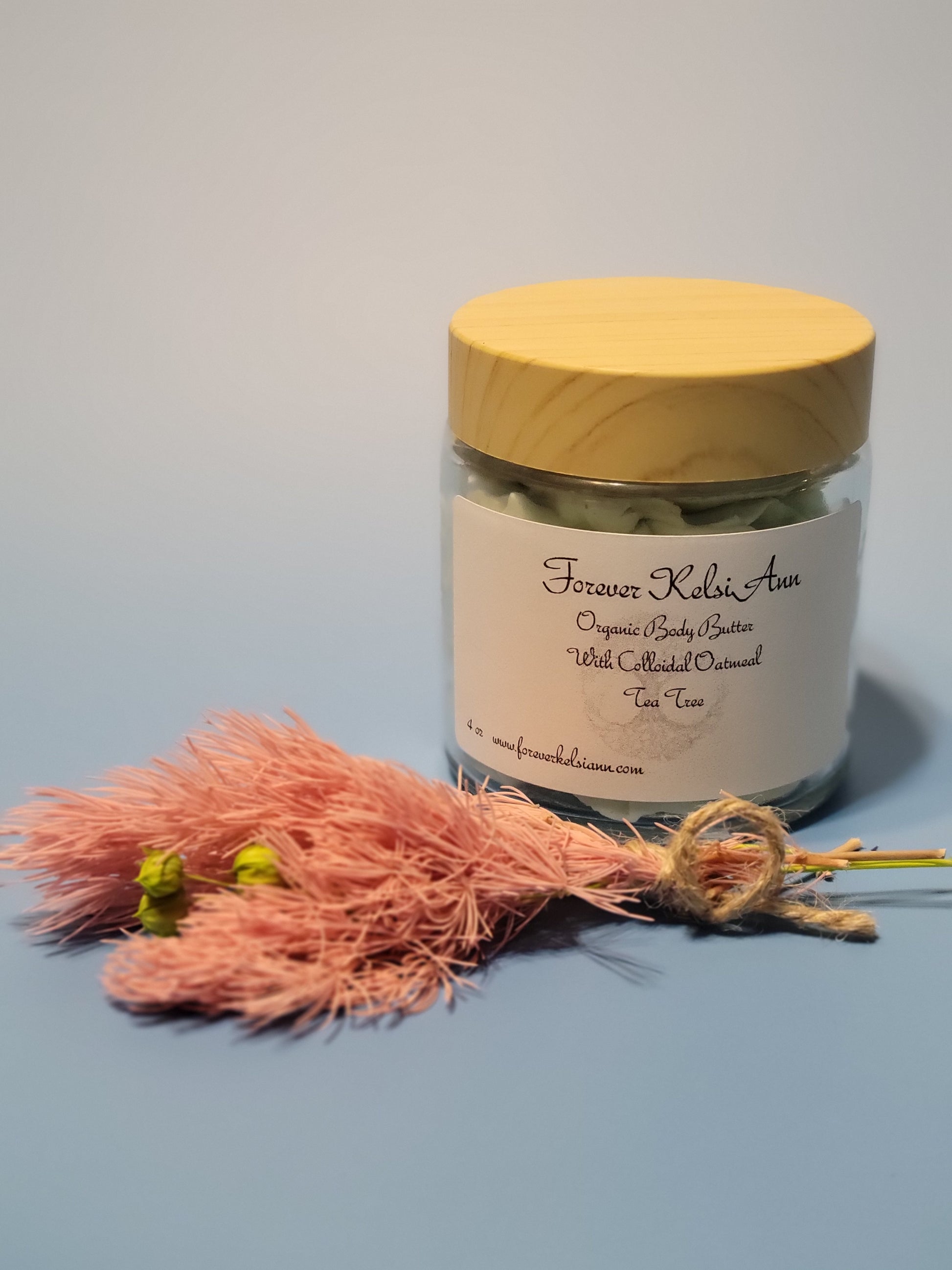 Glass jar of organic Colloidal Oatmeal body butter infused with Tea Tree oil, with dried flowers on light blue background
