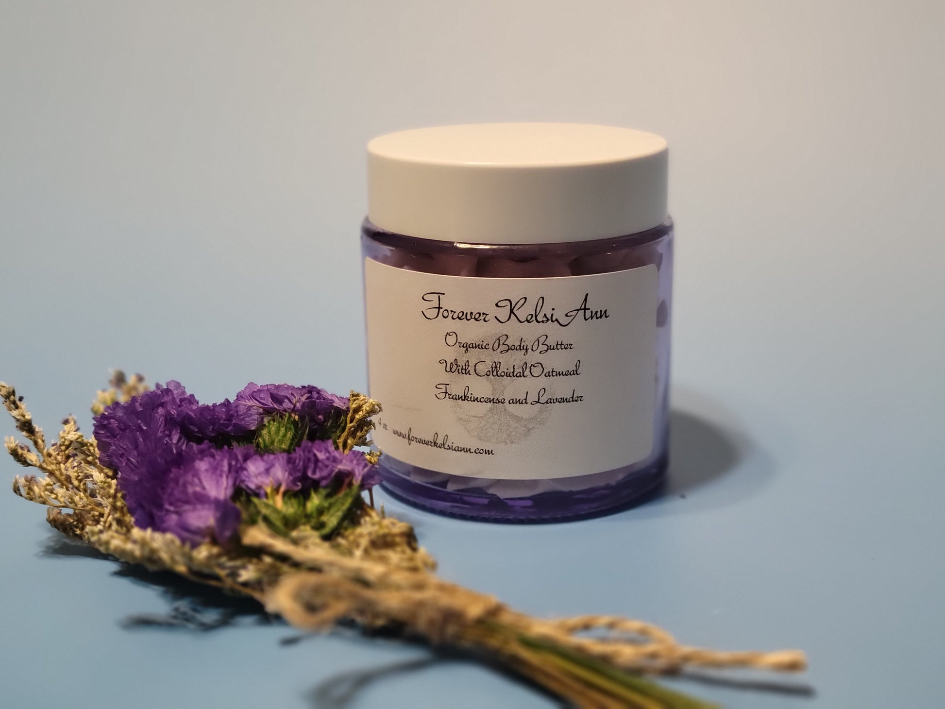 Glass Jar of  Colloidal Oatmeal organic body butter with Frankincense and Lavender on a light blue background with  purple flowers 