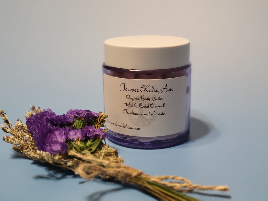 Jar of Forever Kelsi Ann organic Colloidal Oatmeal body butter infused with Lavender and Frankincense, with a bouquet of purple flowers on a light blue background